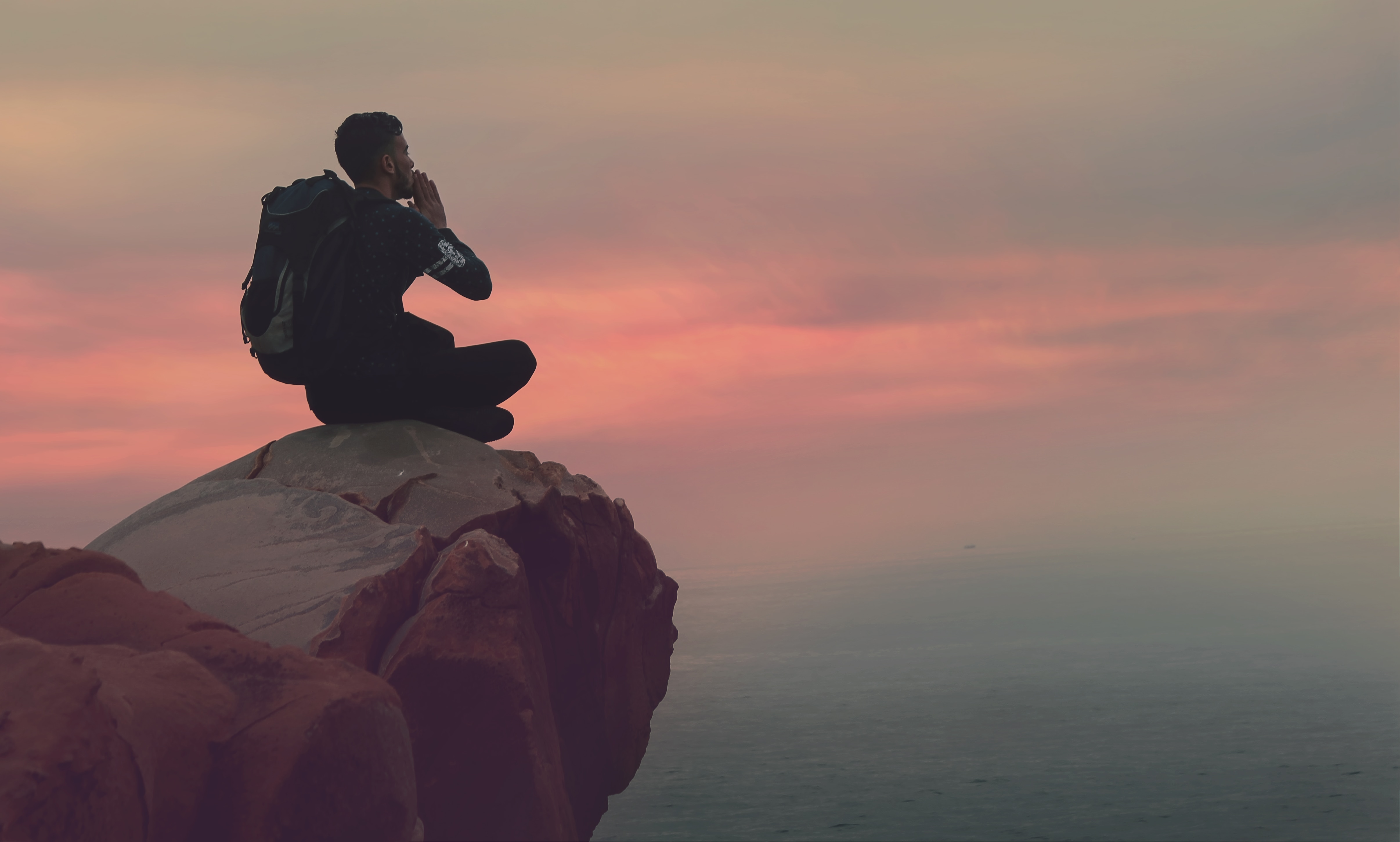 man in black shirt sitting on rock cliff during sunset