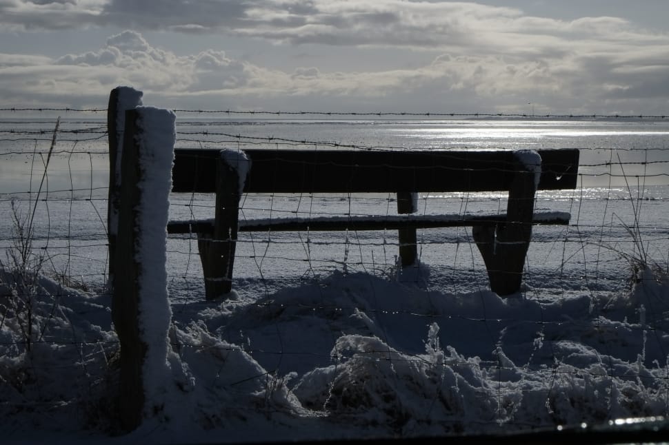 grayscale photo of patio bench in front of wire fence showing beach under white clouds preview