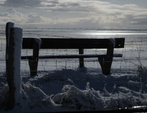 grayscale photo of patio bench in front of wire fence showing beach under white clouds thumbnail
