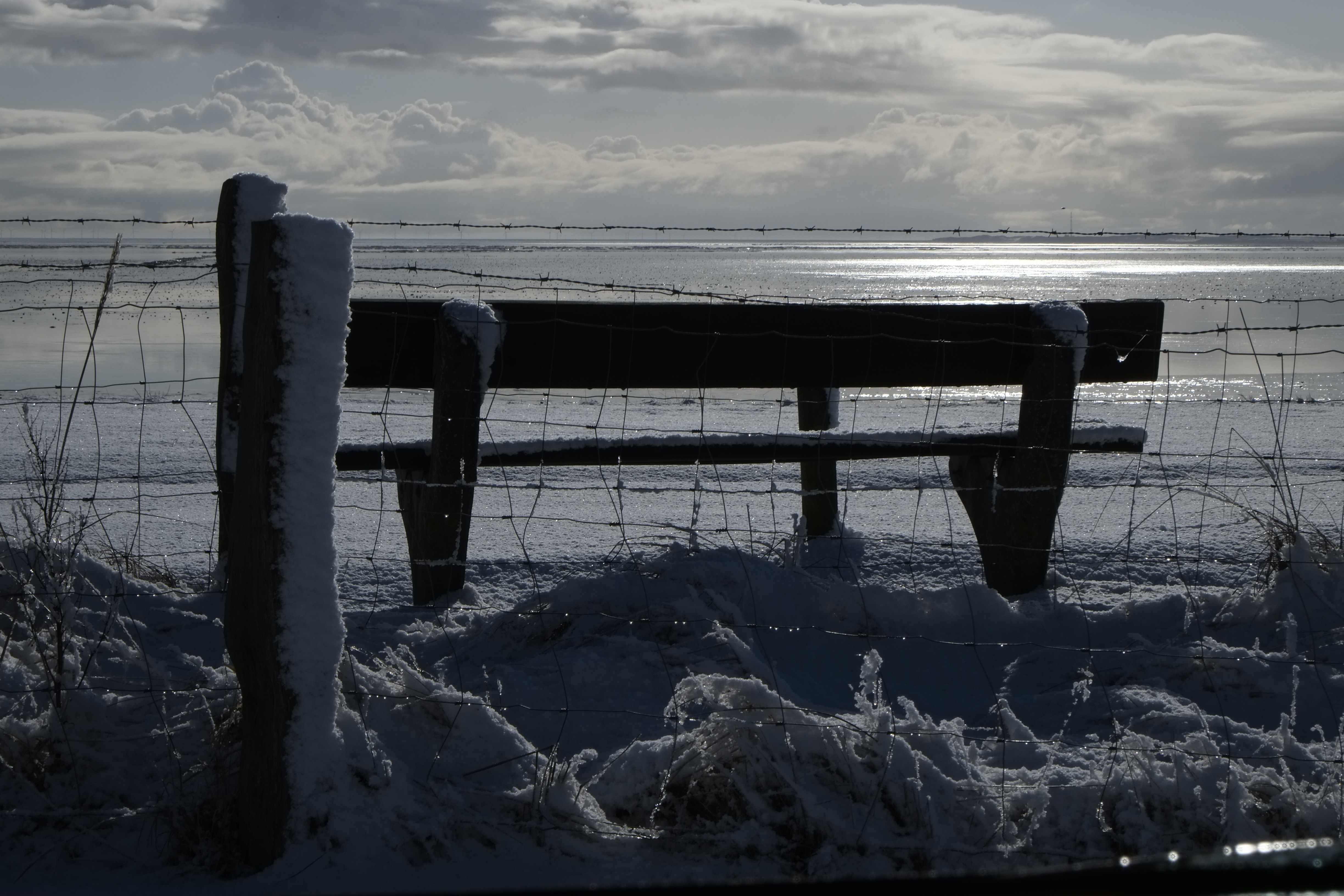 grayscale photo of patio bench in front of wire fence showing beach under white clouds