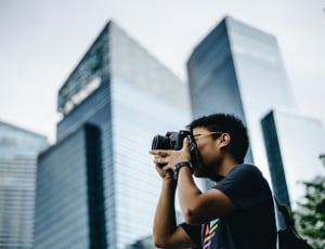 man in black shirt taking photo using black dslr near high rise building at daytime thumbnail