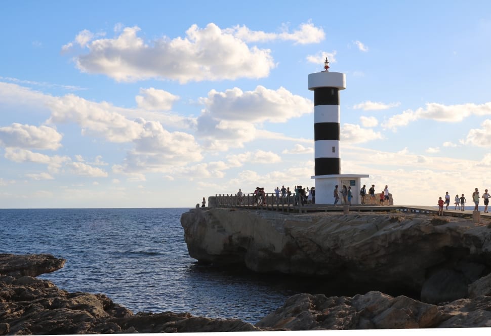 people near white and black concrete lighthouse near blue sea under blue and white cloudy sky during daytime preview