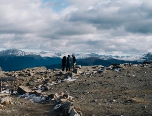 three people standing near cliff overlooking ice-capped mountains under cloudy sky during daytime thumbnail
