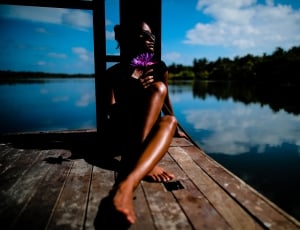 woman wearing sunglasses sitting on brown wooden parquet floor beside body of water during daytime thumbnail