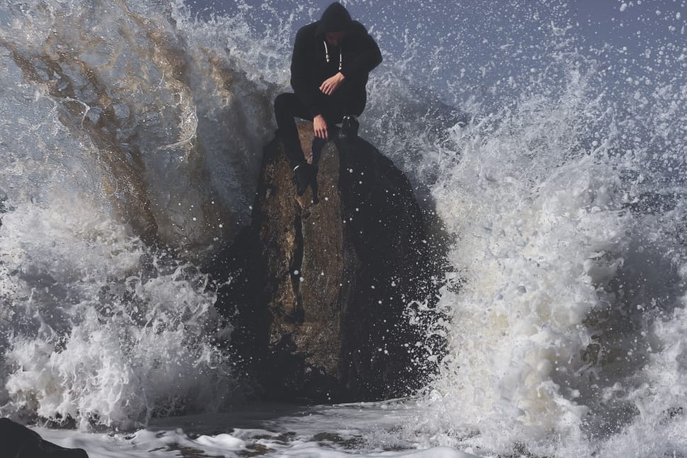 man in black suit sitting on rock formation white hit by big sea waves preview