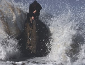 man in black suit sitting on rock formation white hit by big sea waves thumbnail