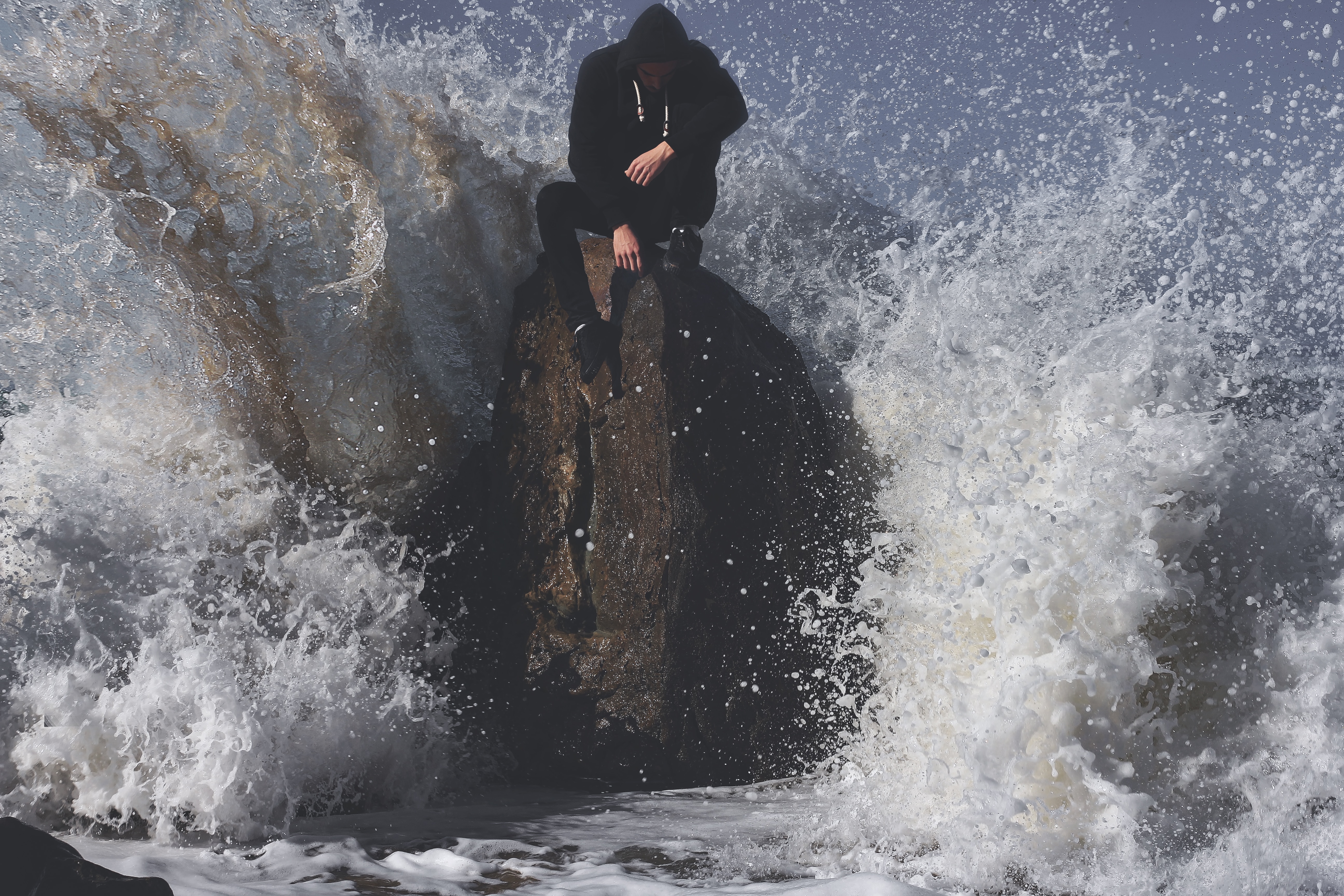 man in black suit sitting on rock formation white hit by big sea waves