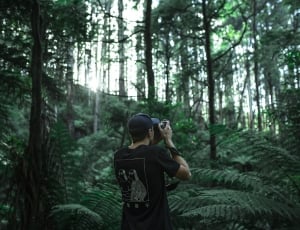 man in black crew neck shirt and black cap holding dslr camera surrounded by green leaf trees at daytime thumbnail